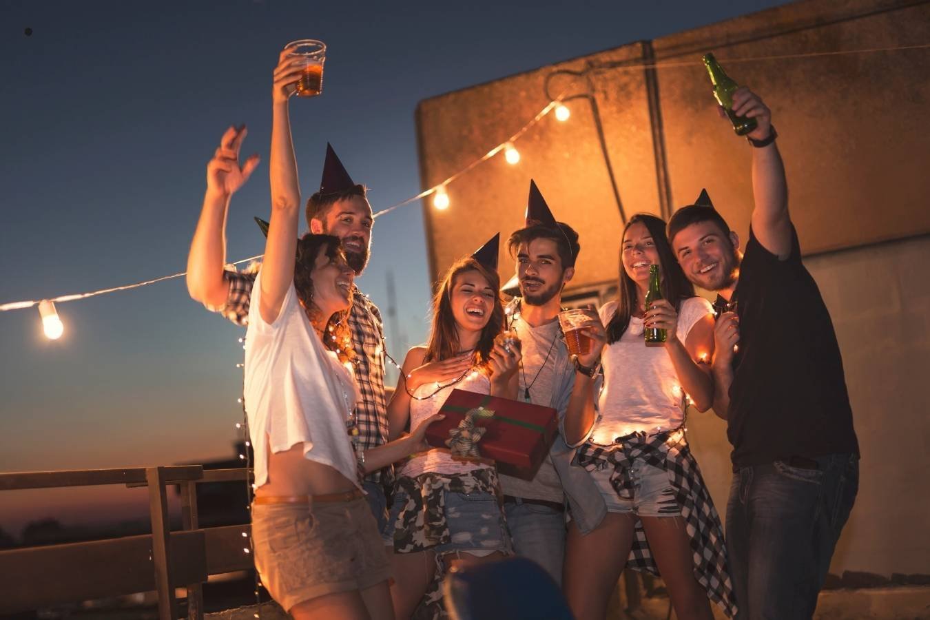 A joyful birthday celebration scene where a man presents a birthday cake with candles to his girlfriend surrounded by friends. Everyone is smiling, holding drinks, and enjoying the moment in a warmly lit room, embodying the essence of heartfelt birthday wishes for a girlfriend.