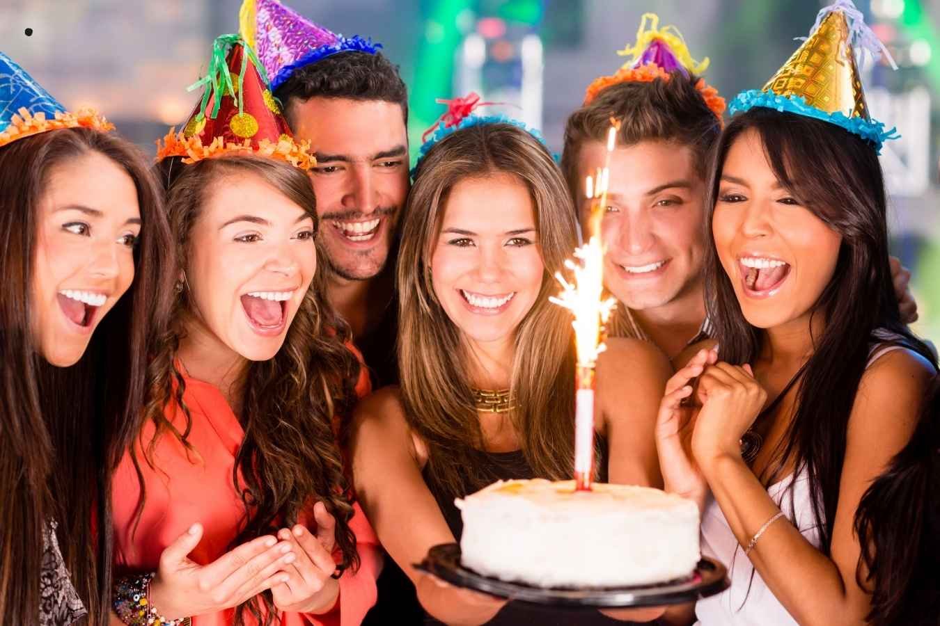 A joyful birthday celebration scene where a man presents a birthday cake with candles to his girlfriend surrounded by friends. Everyone is smiling, holding drinks, and enjoying the moment in a warmly lit room, embodying the essence of heartfelt birthday wishes for a girlfriend.