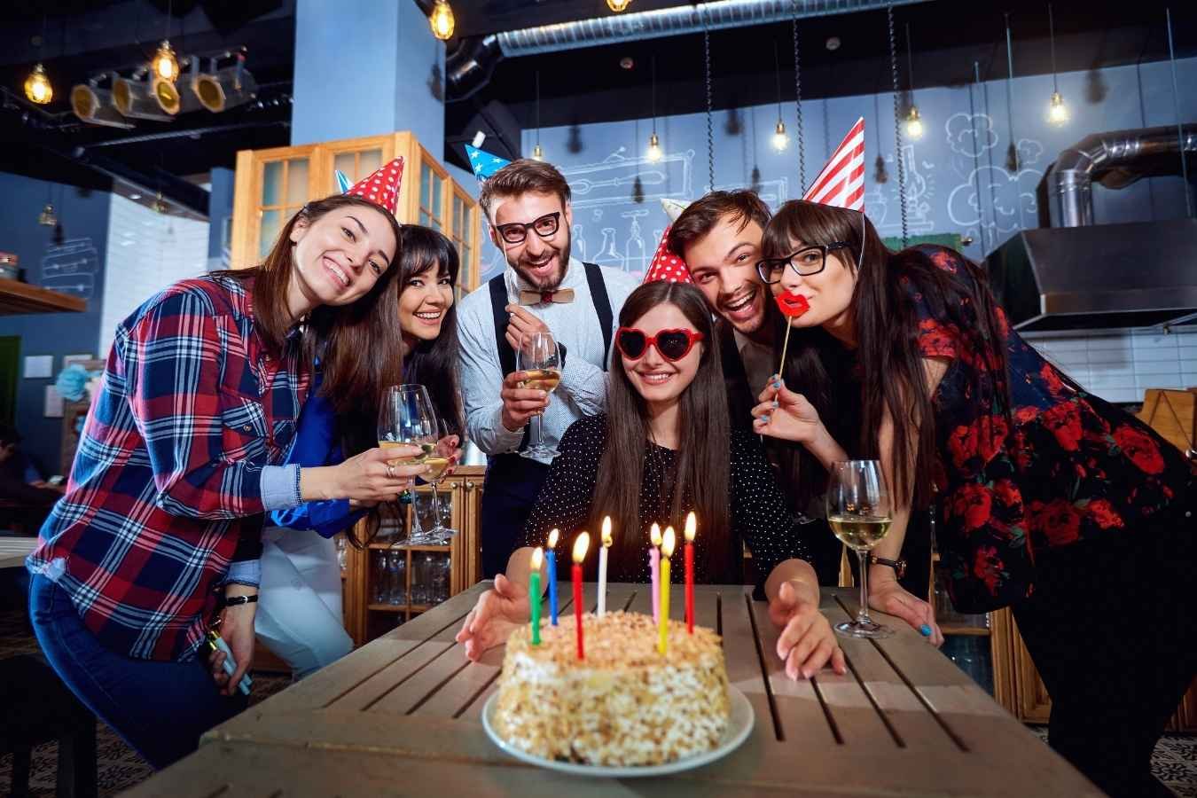 A joyful birthday celebration scene where a man presents a birthday cake with candles to his girlfriend surrounded by friends. Everyone is smiling, holding drinks, and enjoying the moment in a warmly lit room, embodying the essence of heartfelt birthday wishes for a girlfriend.