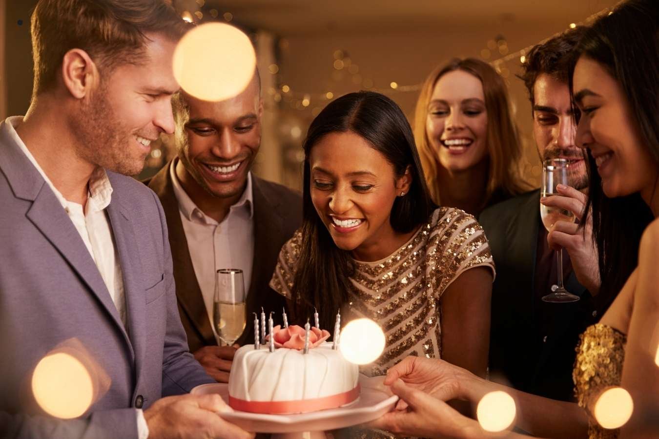 A joyful birthday celebration scene where a man presents a birthday cake with candles to his girlfriend surrounded by friends. Everyone is smiling, holding drinks, and enjoying the moment in a warmly lit room, embodying the essence of heartfelt birthday wishes for a girlfriend.