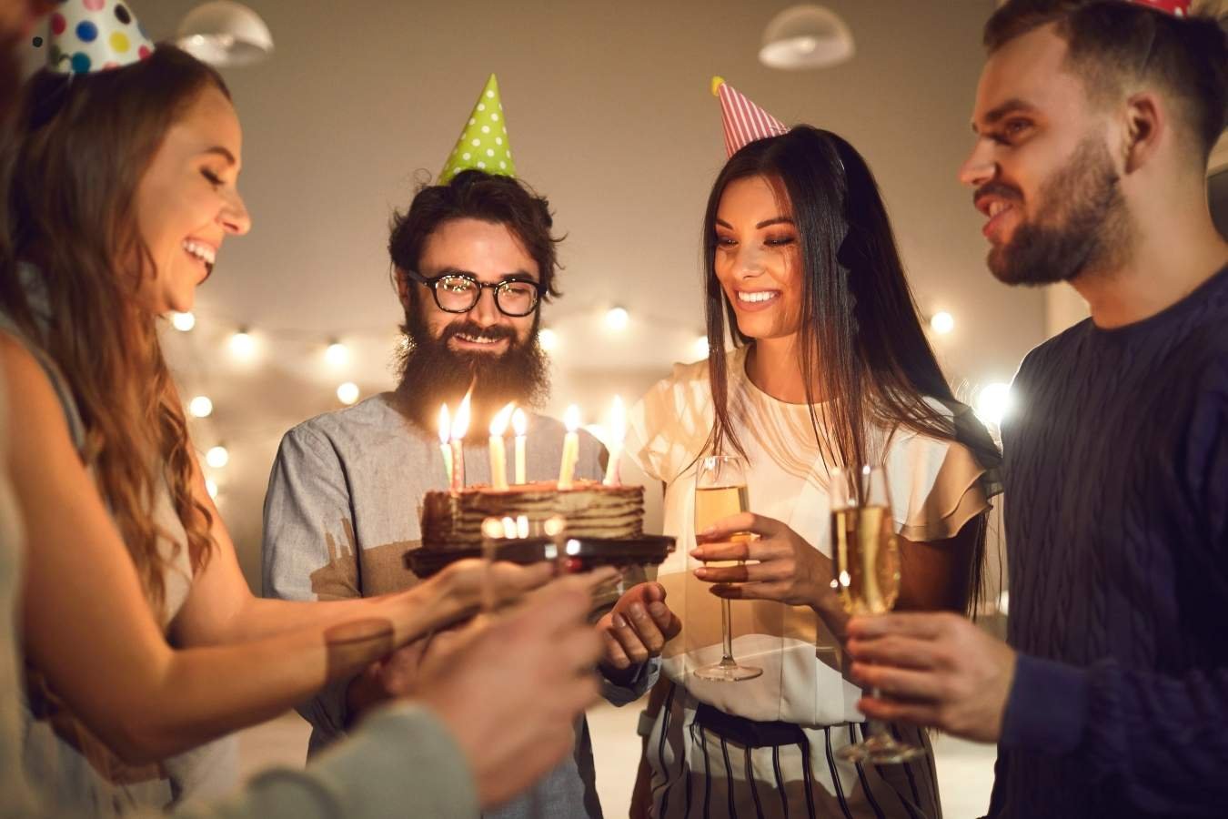A joyful birthday celebration scene where a man presents a birthday cake with candles to his girlfriend surrounded by friends. Everyone is smiling, holding drinks, and enjoying the moment in a warmly lit room, embodying the essence of heartfelt birthday wishes for a girlfriend.