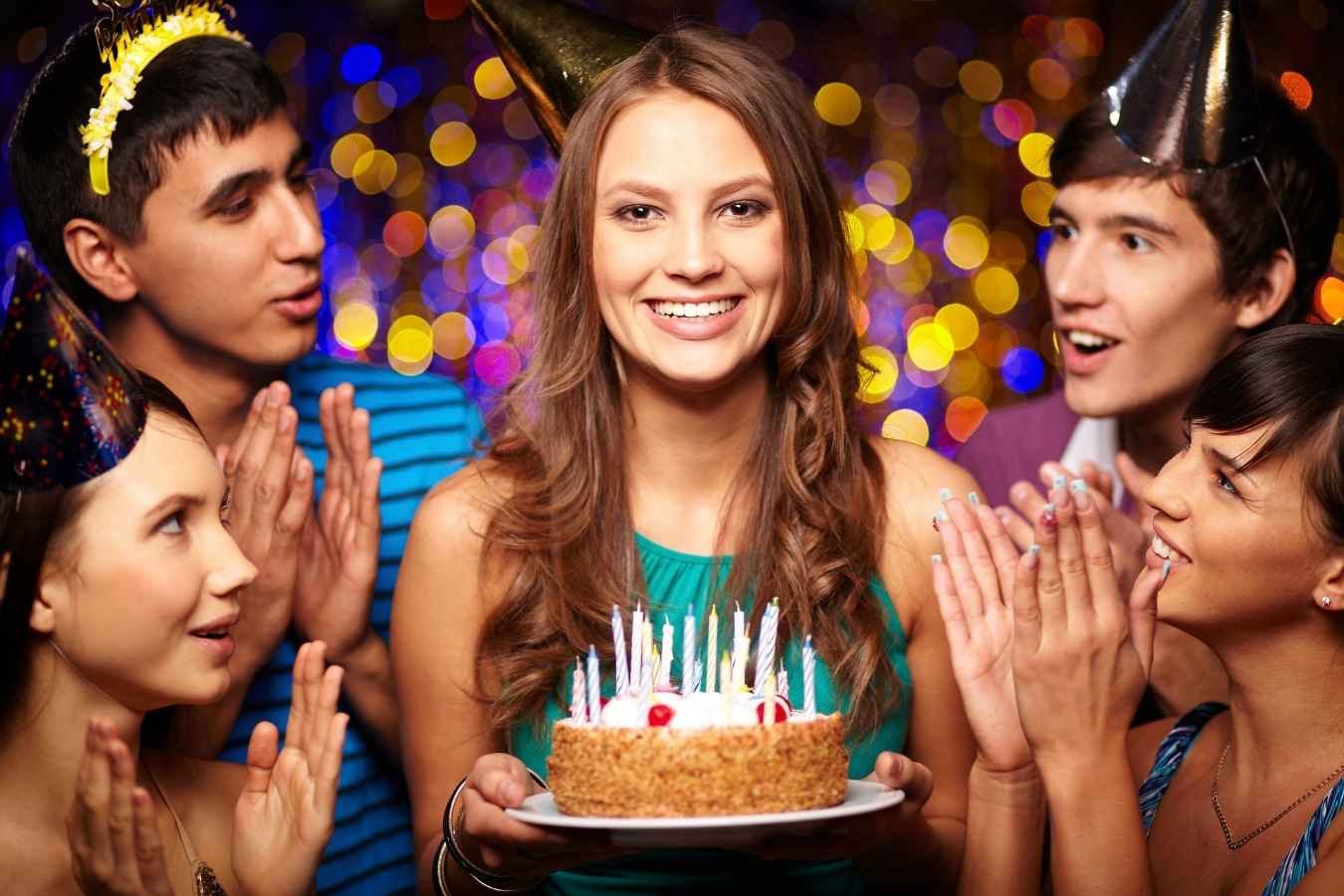 A joyful birthday celebration scene where a man presents a birthday cake with candles to his girlfriend surrounded by friends. Everyone is smiling, holding drinks, and enjoying the moment in a warmly lit room, embodying the essence of heartfelt birthday wishes for a girlfriend.