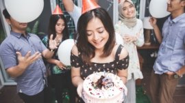 A joyful birthday celebration scene where a man presents a birthday cake with candles to his girlfriend surrounded by friends. Everyone is smiling, holding drinks, and enjoying the moment in a warmly lit room, embodying the essence of heartfelt birthday wishes for a girlfriend.