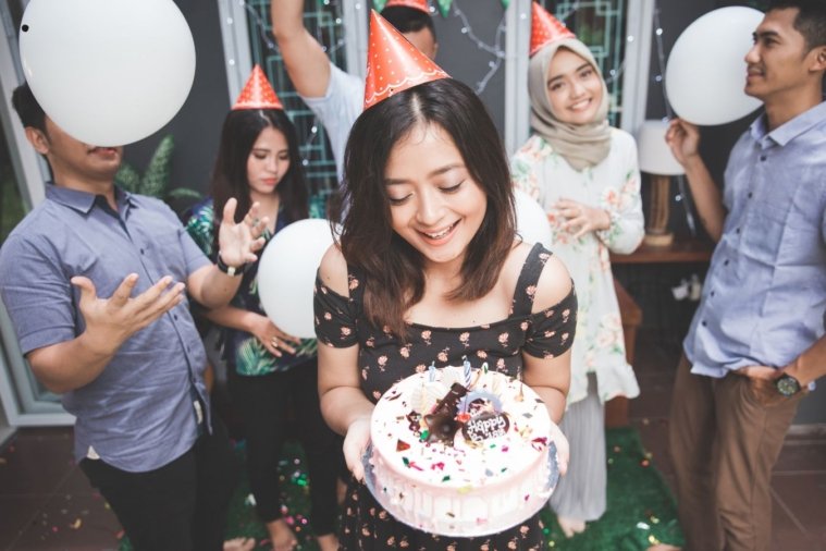 A joyful birthday celebration scene where a man presents a birthday cake with candles to his girlfriend surrounded by friends. Everyone is smiling, holding drinks, and enjoying the moment in a warmly lit room, embodying the essence of heartfelt birthday wishes for a girlfriend.