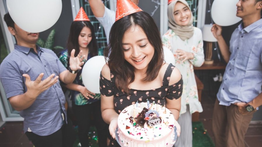 A joyful birthday celebration scene where a man presents a birthday cake with candles to his girlfriend surrounded by friends. Everyone is smiling, holding drinks, and enjoying the moment in a warmly lit room, embodying the essence of heartfelt birthday wishes for a girlfriend.