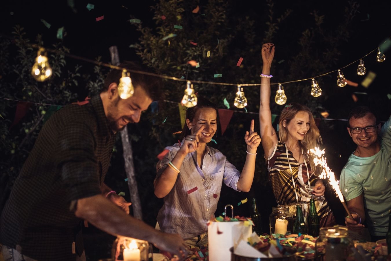 A joyful birthday celebration scene where a man presents a birthday cake with candles to his girlfriend surrounded by friends. Everyone is smiling, holding drinks, and enjoying the moment in a warmly lit room, embodying the essence of heartfelt birthday wishes for a girlfriend.